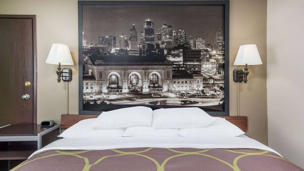 Een hotelkamer met een bed, twee bedlampen, een stadsgezicht in een lijst aan de muur, en een houten deur.