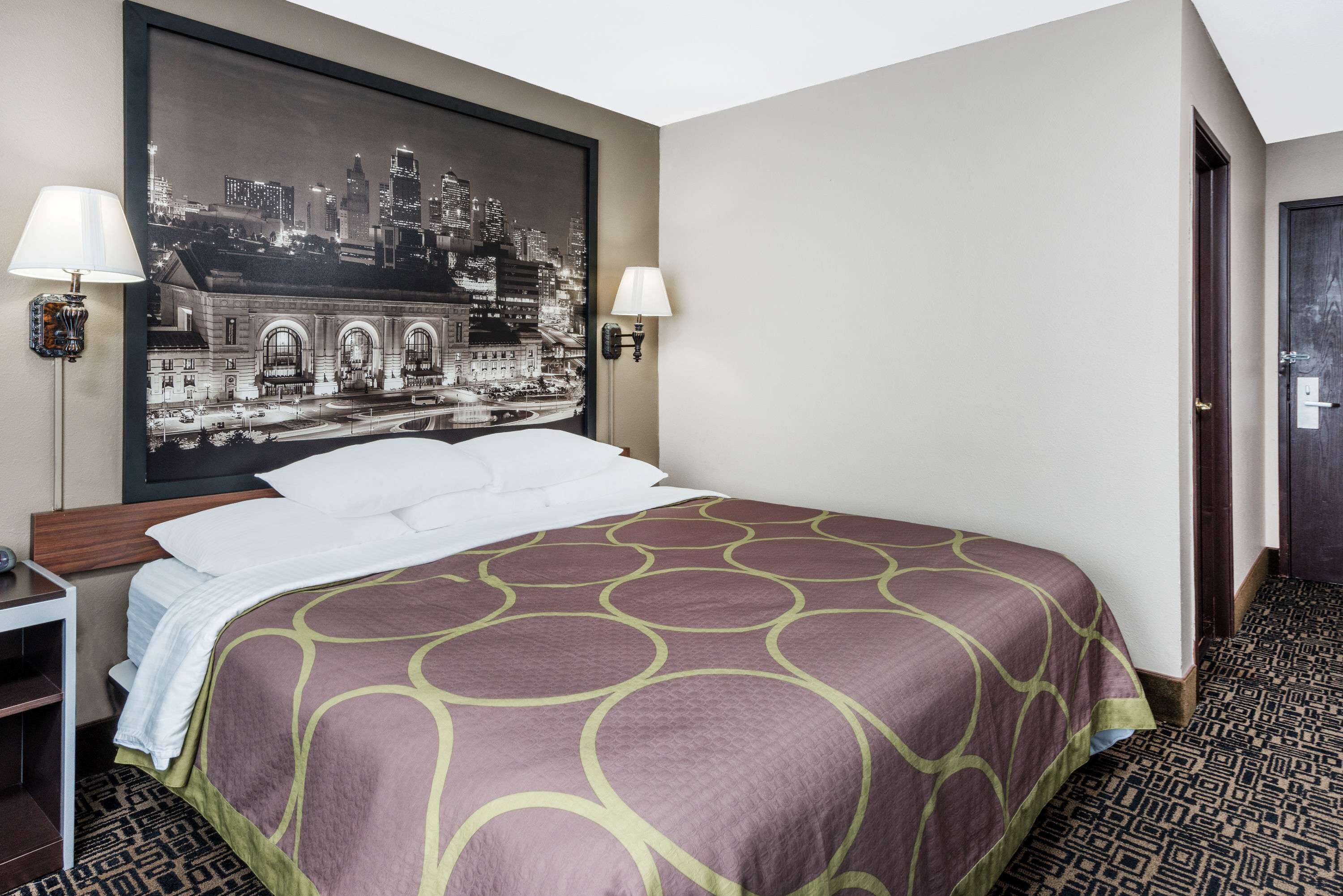 A hotel room with a large bed, two bedside lamps, a cityscape painting on the wall, and a patterned carpet.