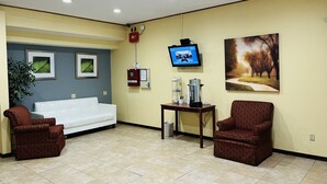 A waiting area with a white sofa, two patterned chairs, a coffee station, a TV, framed pictures, and a plant.
