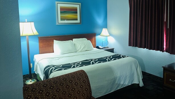 A hotel room with a bed, two bedside lamps, a nightstand, a chair, a painting on the wall, and a window with curtains.