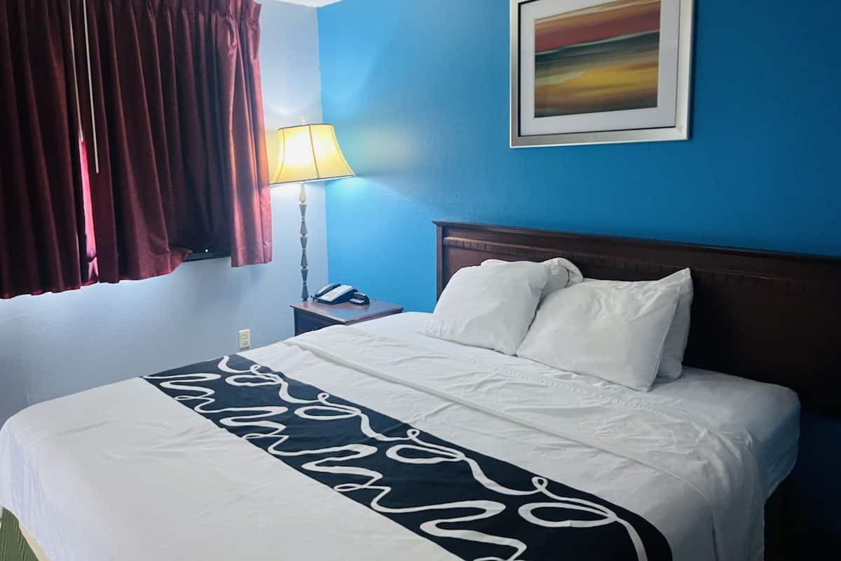 A hotel room with a bed, a nightstand, a lamp, a painting on the wall, and a window with curtains.