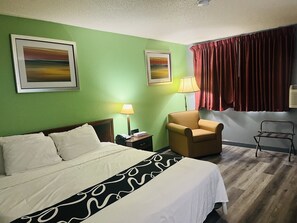 A hotel room with a bed, a chair, a nightstand, a lamp, and two paintings on the wall.