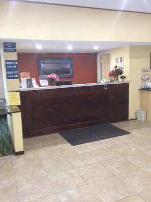 A reception area with a wooden reception desk, a flat-screen TV mounted on the wall, a check-in/out sign, and a small potted plant with pink flowers.