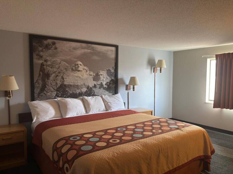 Habitación de hotel con cama, un cuadro grande enmarcado en la pared, dos lámparas de noche, una ventana con cortinas y piso alfombrado.