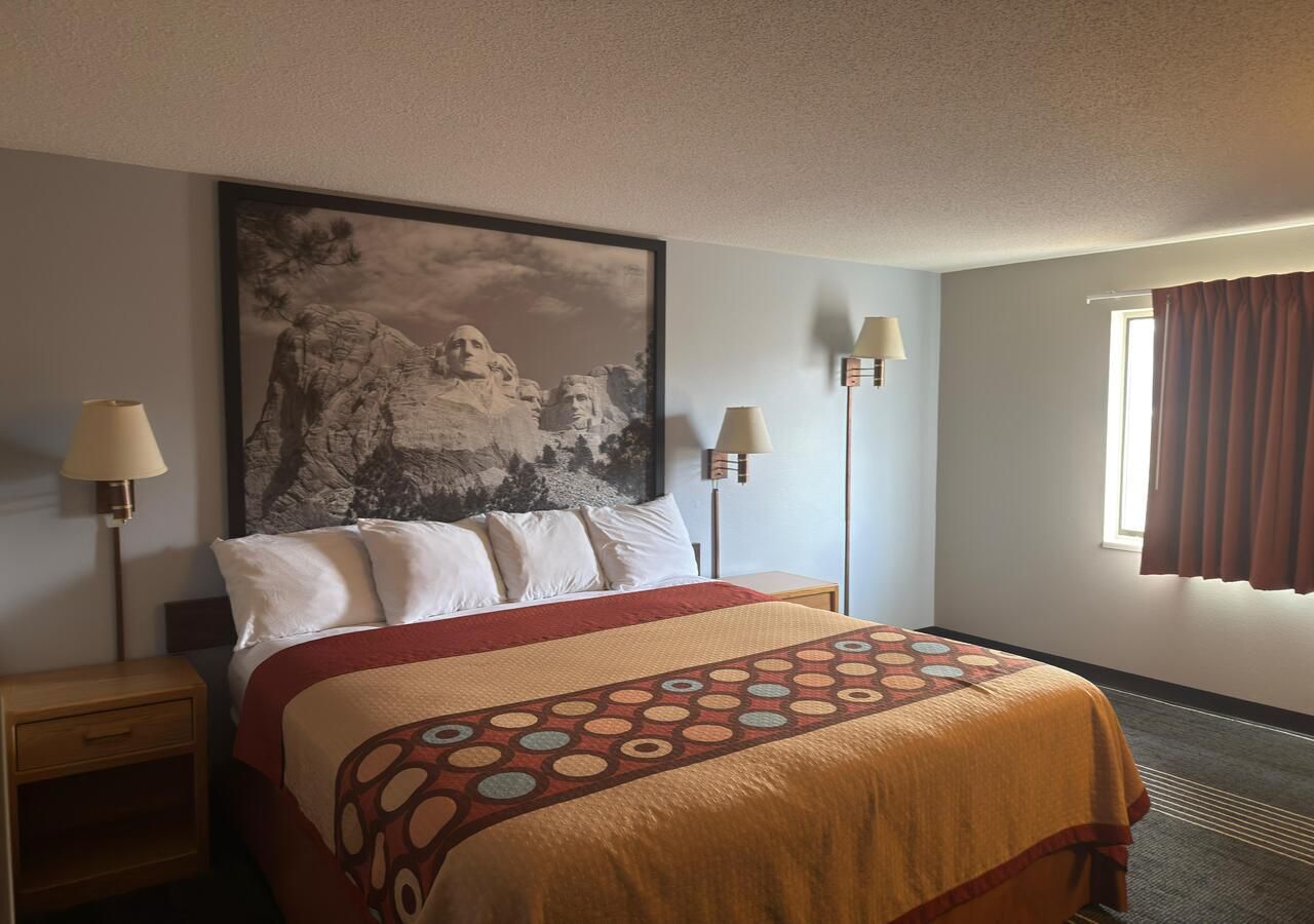 A hotel room with a bed, two bedside lamps, a large framed picture on the wall, and a window with curtains.