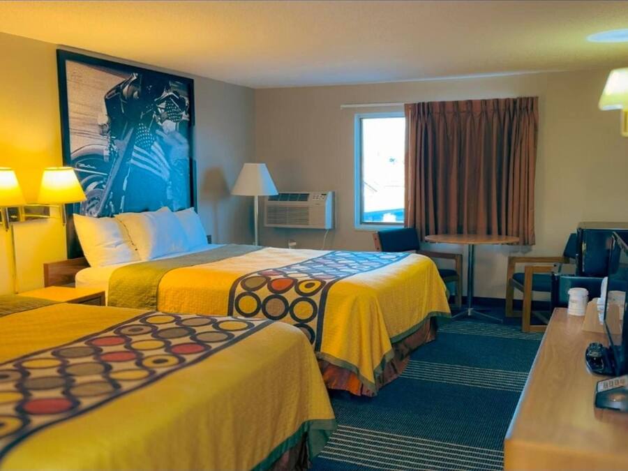 A hotel room with two beds, a desk, a chair, a lamp, an air conditioner, and a window with curtains.