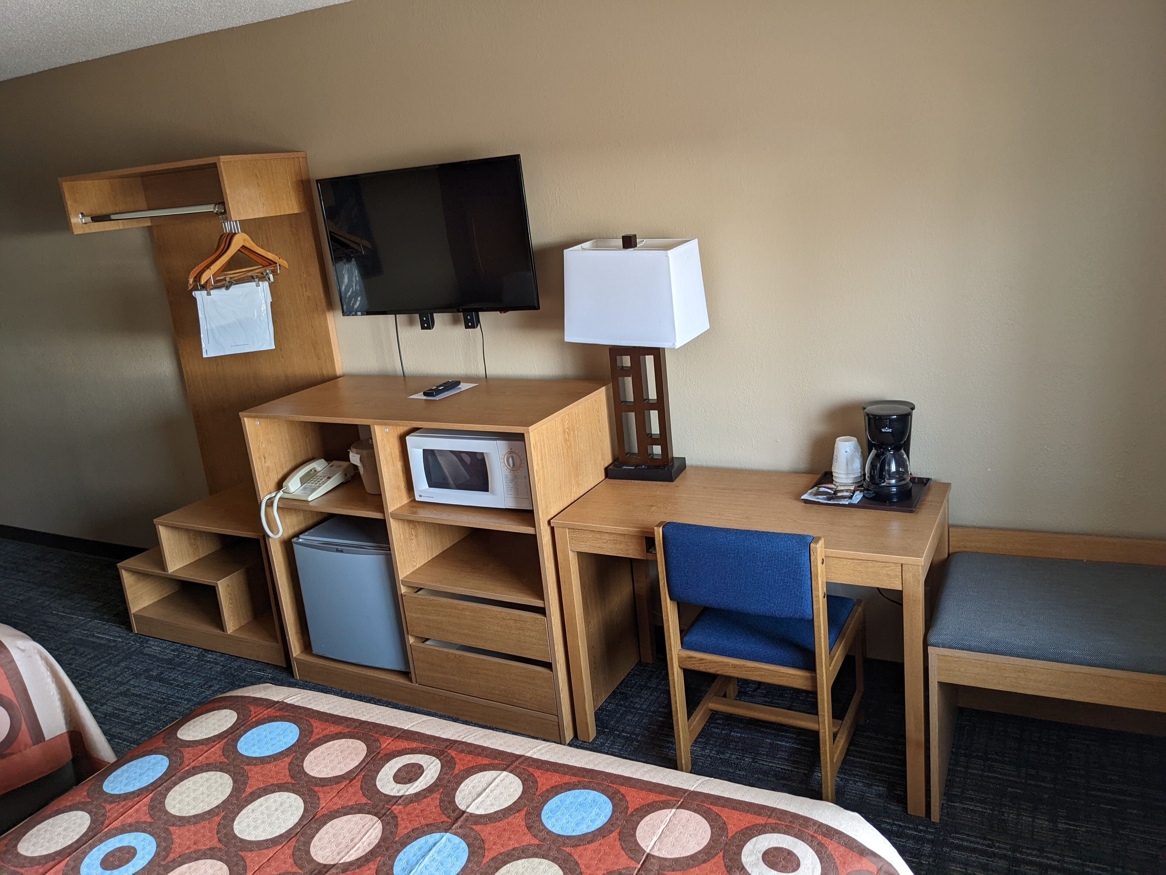 Habitació d'hotel amb escriptori de fusta, televisió penjada a la paret, microones, cafetera i una cadira blava.
