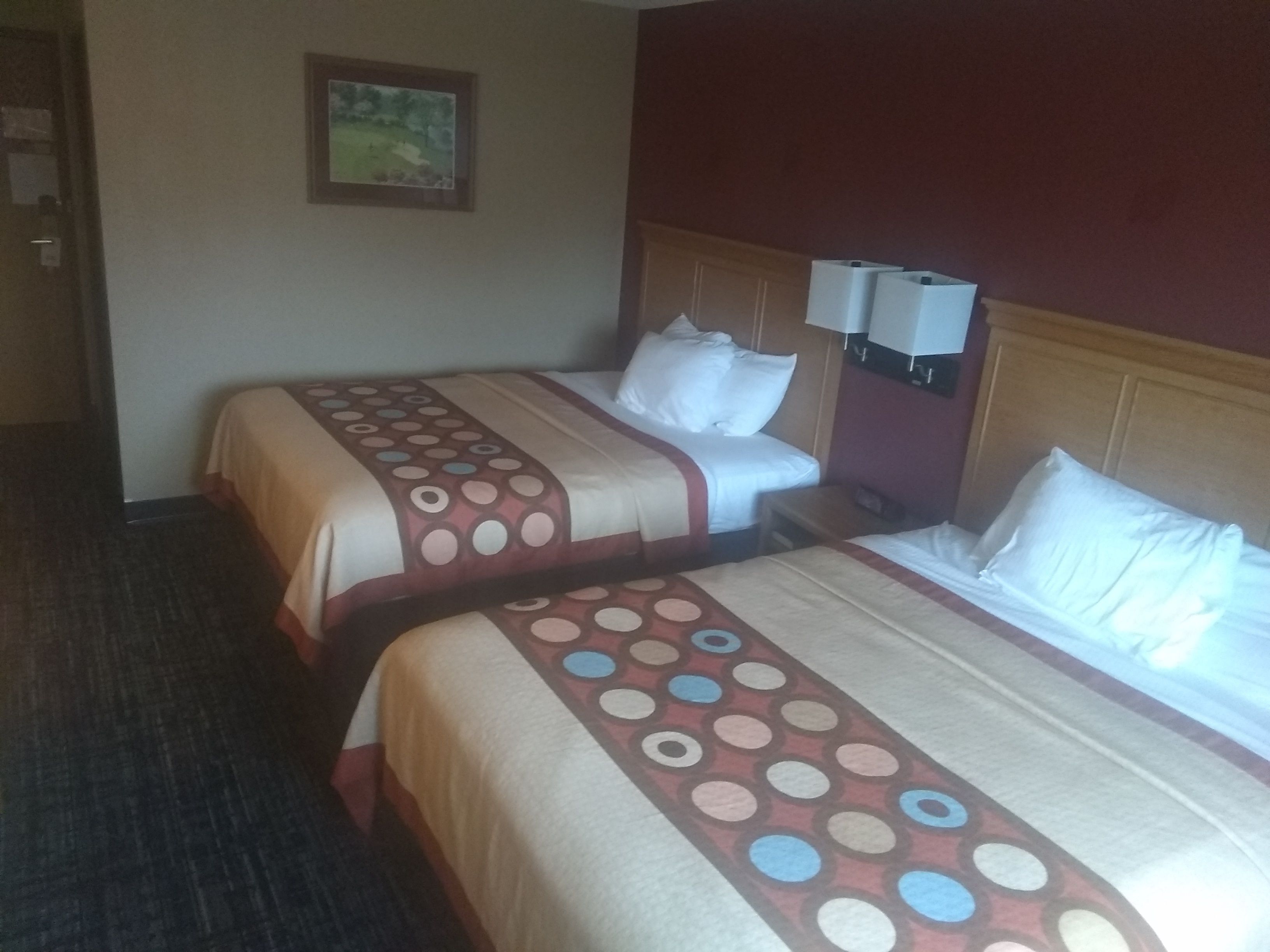 Une chambre d’hôtel avec deux lits, une tête de lit, une lampe fixée au mur et un tableau encadré.