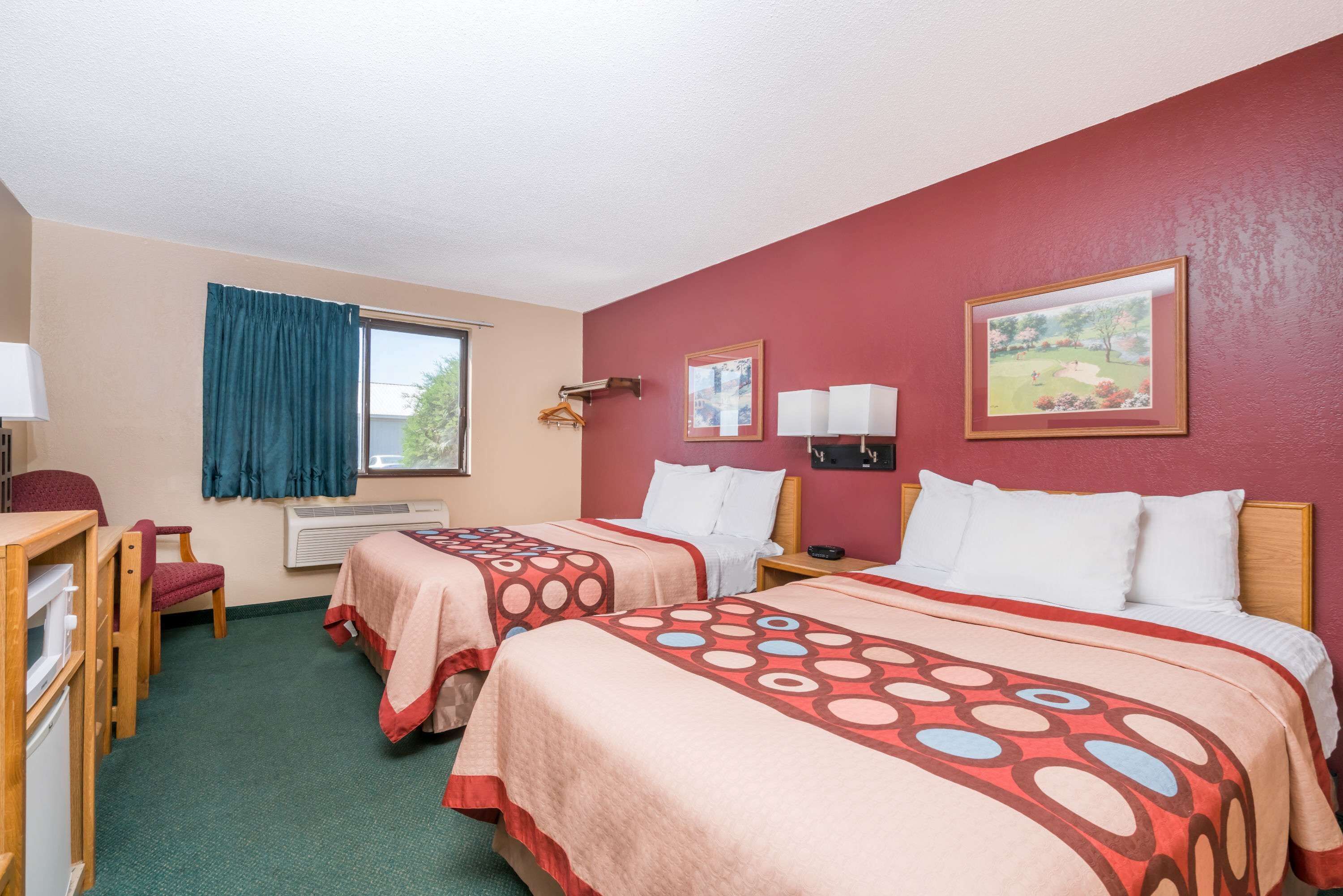 Ein Hotelzimmer mit zwei Betten, einem Schreibtisch und einem Stuhl. Zwei Wandleuchten und ein Fenster mit Vorhang sind vorhanden.