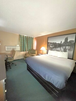 Quarto de hotel com cama, escrivaninha, ar condicionado, cadeira, luminária e uma pintura de paisagem urbana na parede.