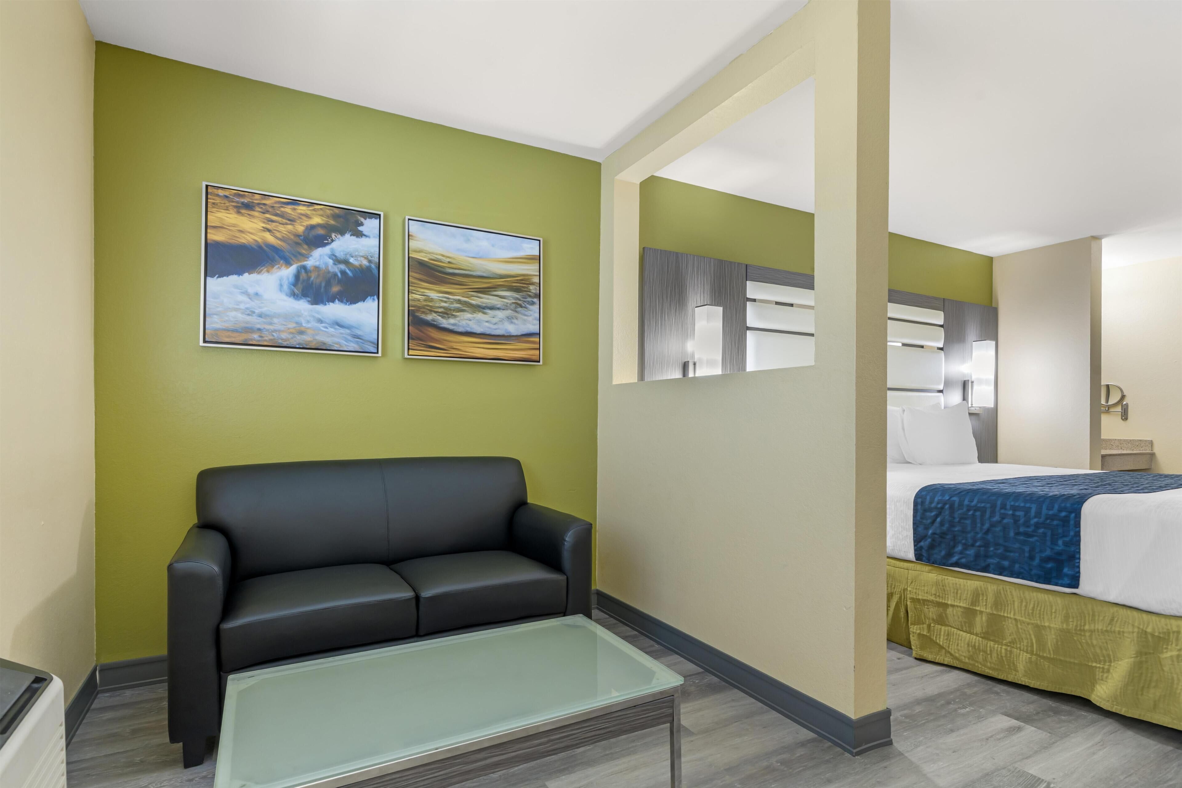 A hotel room with a green wall, a black leather sofa, a glass-top coffee table, a bed with a blue blanket, and two framed pictures.