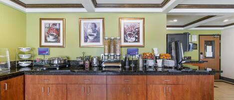 A hotel kitchen with a large island, wooden cabinets, and various appliances. There are framed pictures on the wall and a sign for the coffee station.