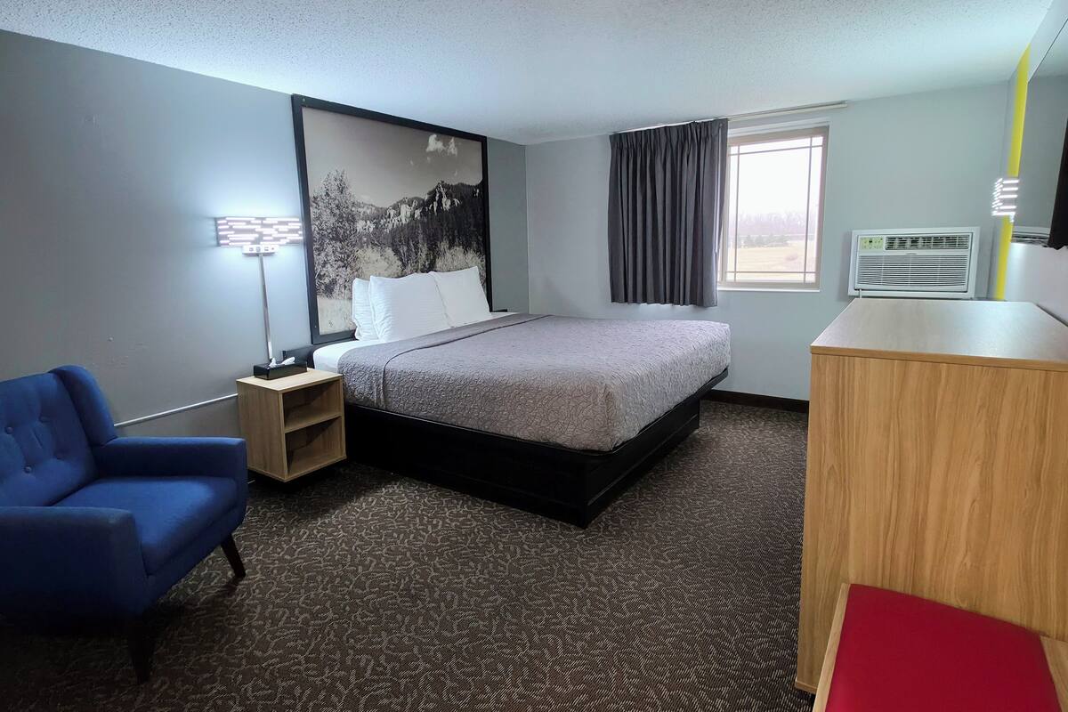 A hotel room with a bed, a desk, a chair, a lamp, a window with curtains, and an air conditioner.