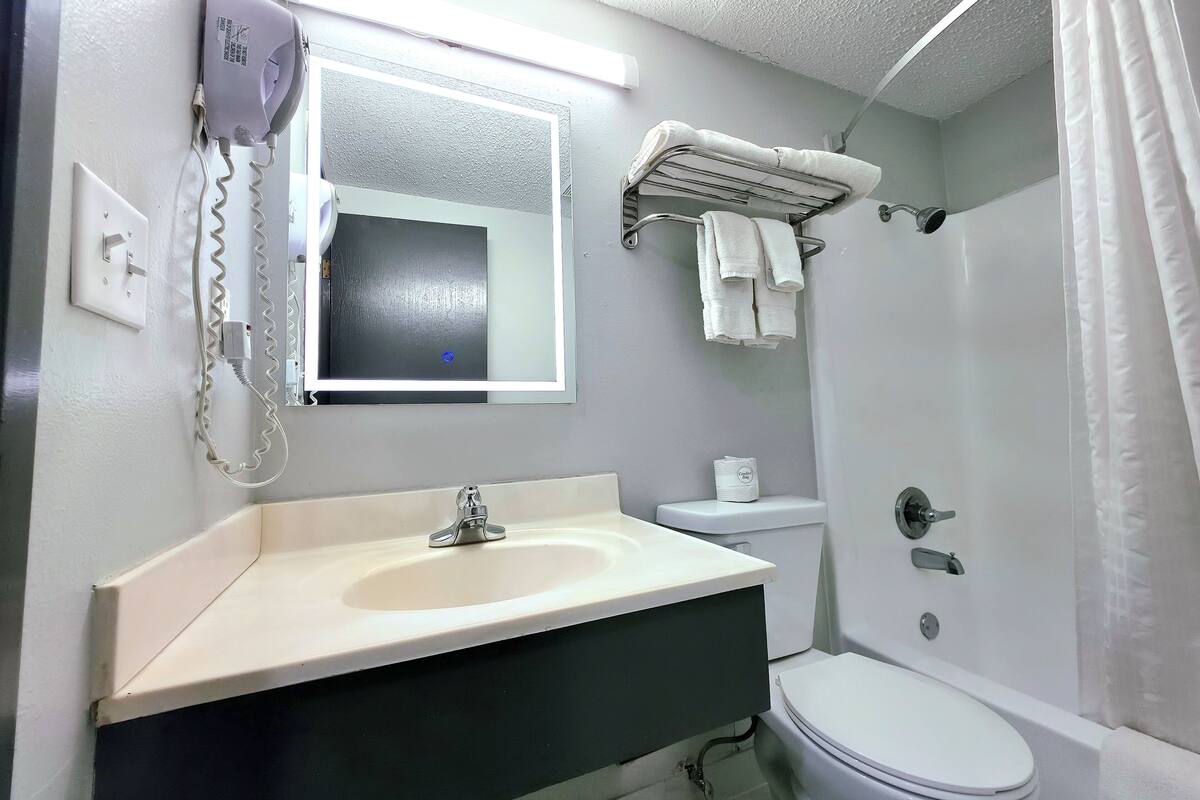 A bathroom with a white sink, mirror, and toilet. There is a hairdryer on the wall, a towel rack above the toilet, and a shower curtain.