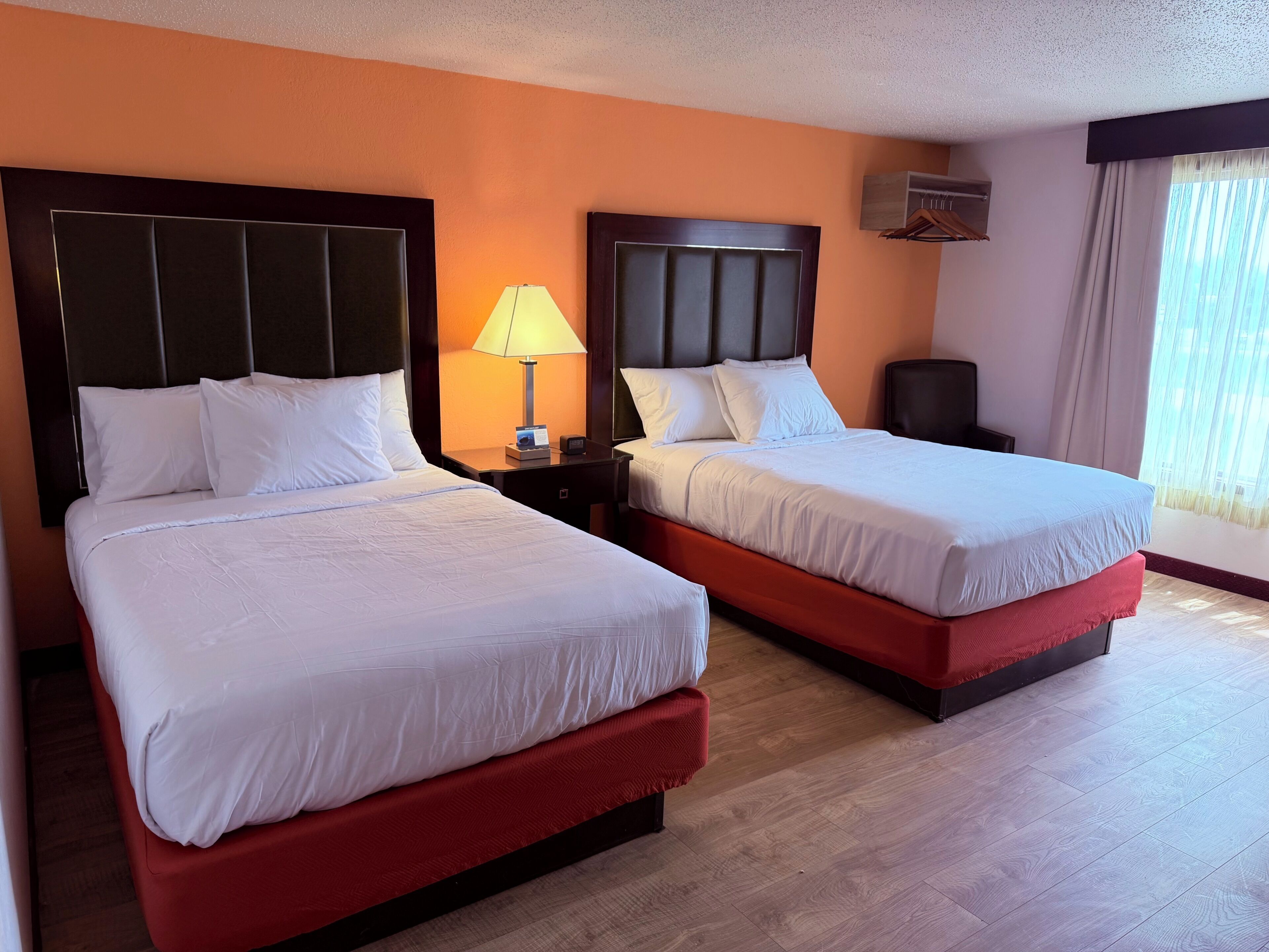 A hotel room with two beds, a nightstand with a lamp, a wall-mounted air conditioner, and a window with curtains.