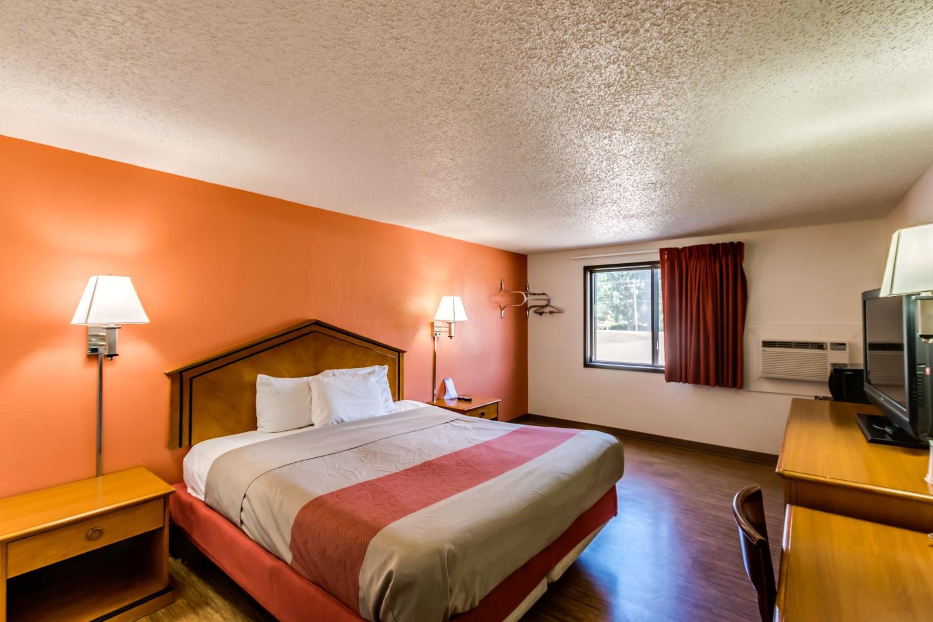 A hotel room with a bed, two bedside lamps, a desk with a television, and a window with curtains.