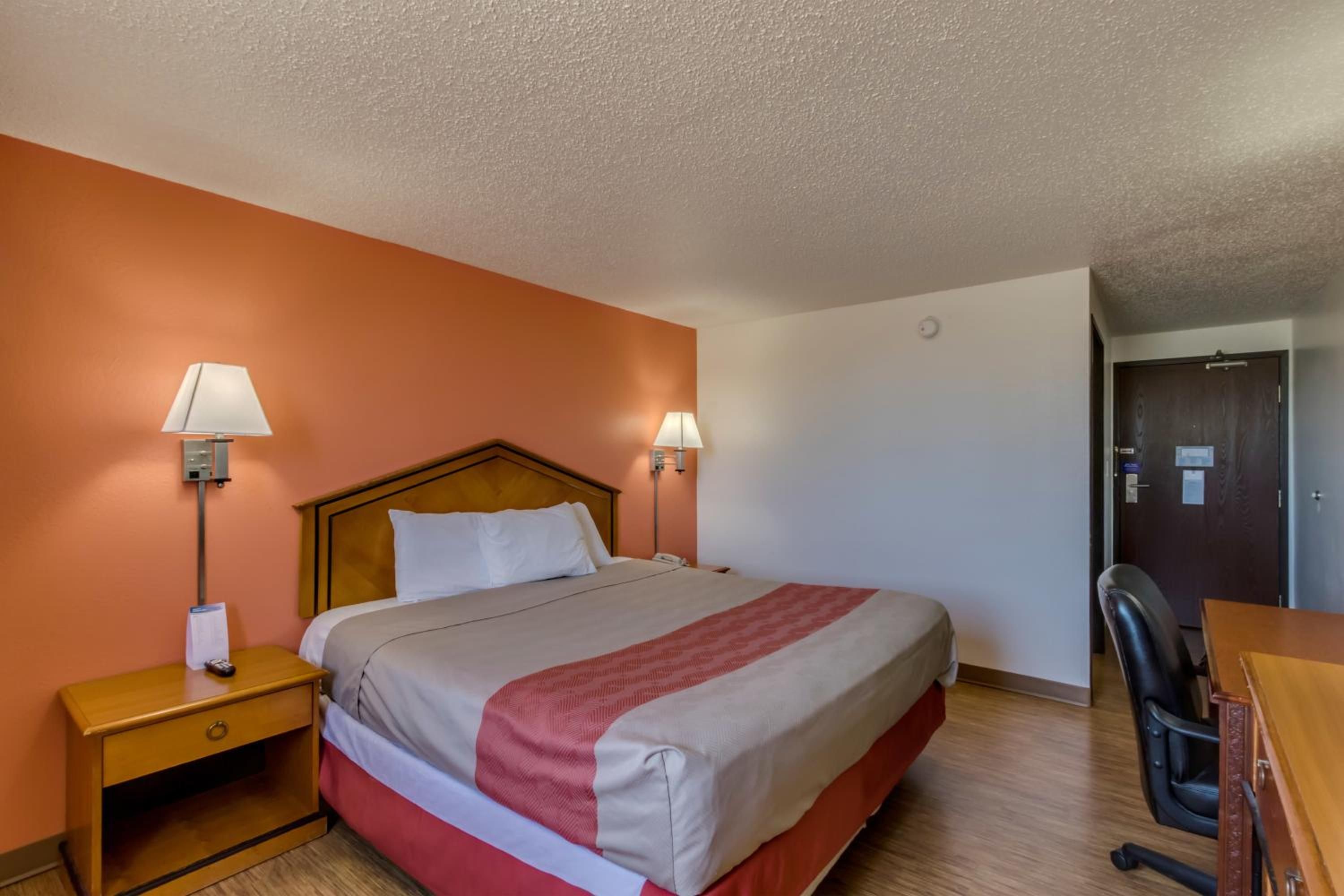 A hotel room with a bed, two bedside lamps, a wooden nightstand, a desk, and a chair.