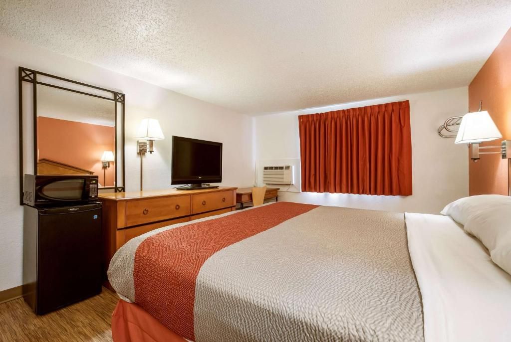 Habitación de hotel con cama, televisor, escritorio con lámpara, microondas y refrigerador.
