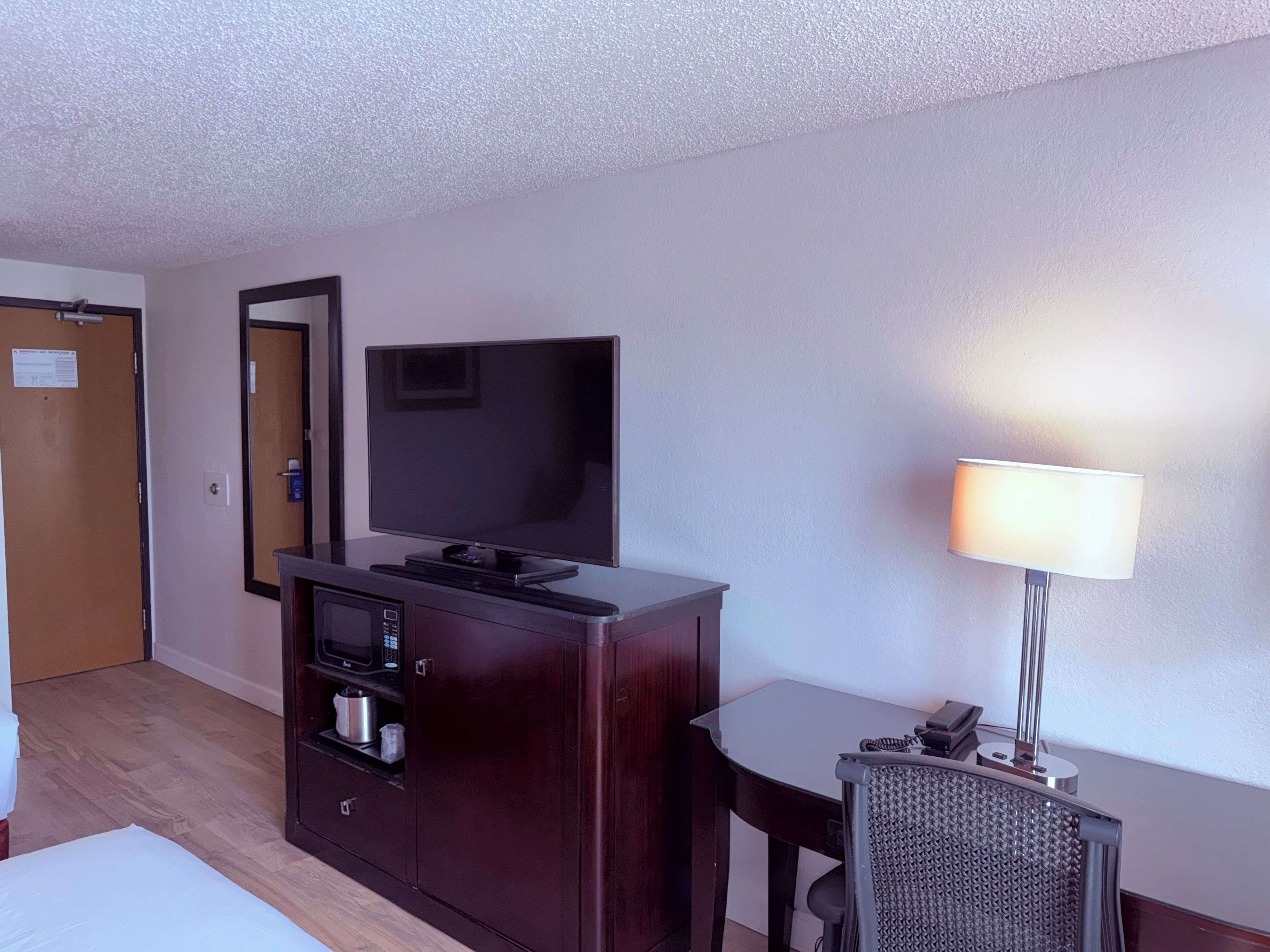 A hotel room with a flat-screen TV on a wooden cabinet, a desk with a lamp, and a chair.