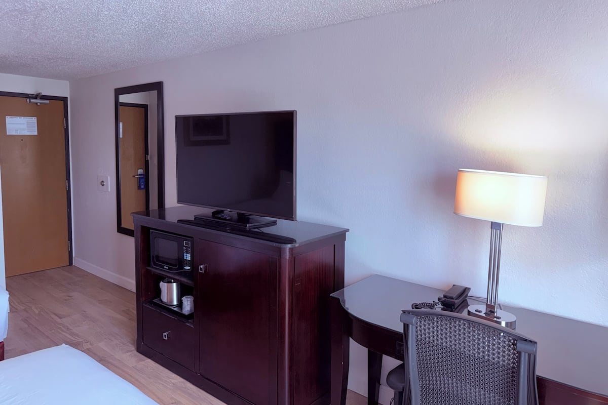 A hotel room with a flat-screen TV on a wooden cabinet, a desk with a lamp, and a chair.
