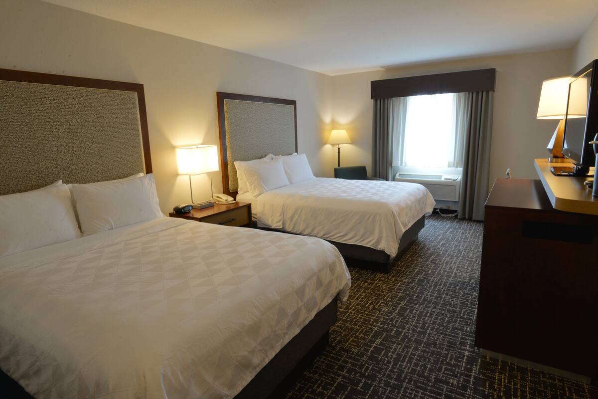 A hotel room with two beds, a desk, a chair, a lamp, and a large window with curtains.