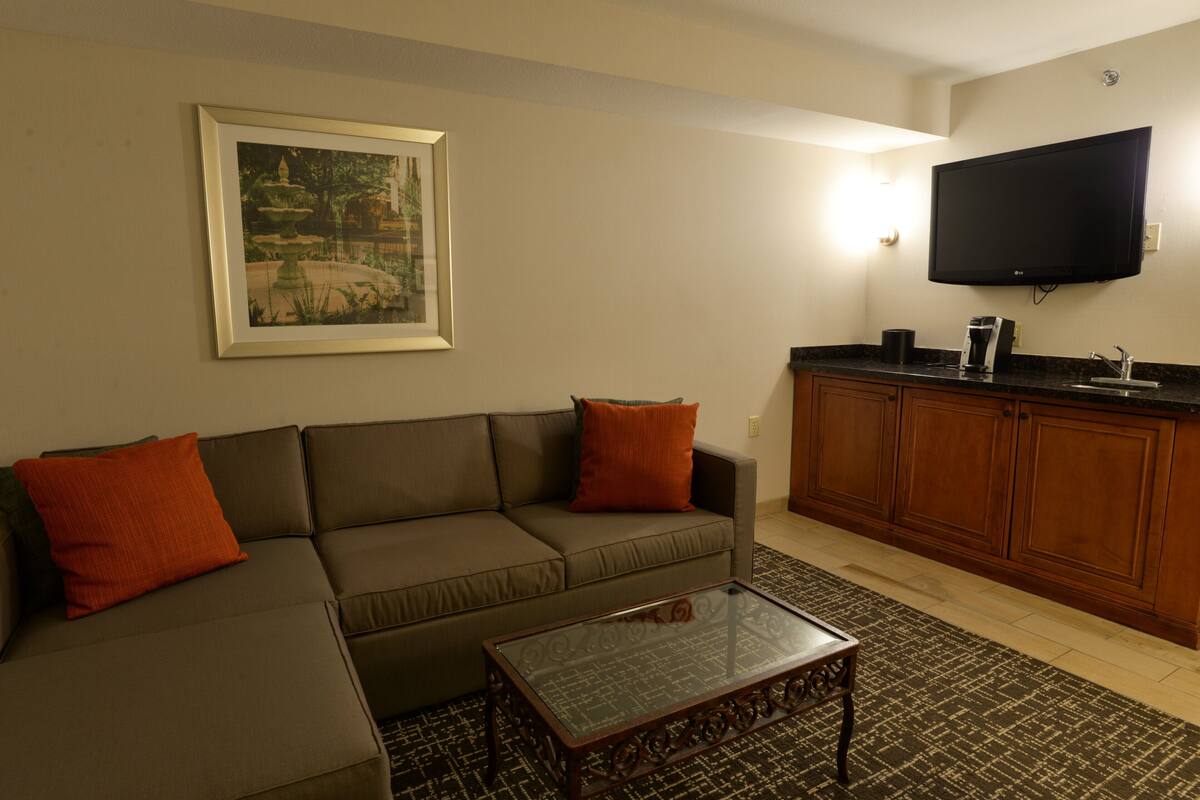 A living room with a sectional sofa, a coffee table, a flat-screen TV mounted above the kitchen counter, and a framed picture on the wall.