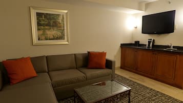 A living room with a sectional sofa, a coffee table, a flat-screen TV mounted above the kitchen counter, and a framed picture on the wall.