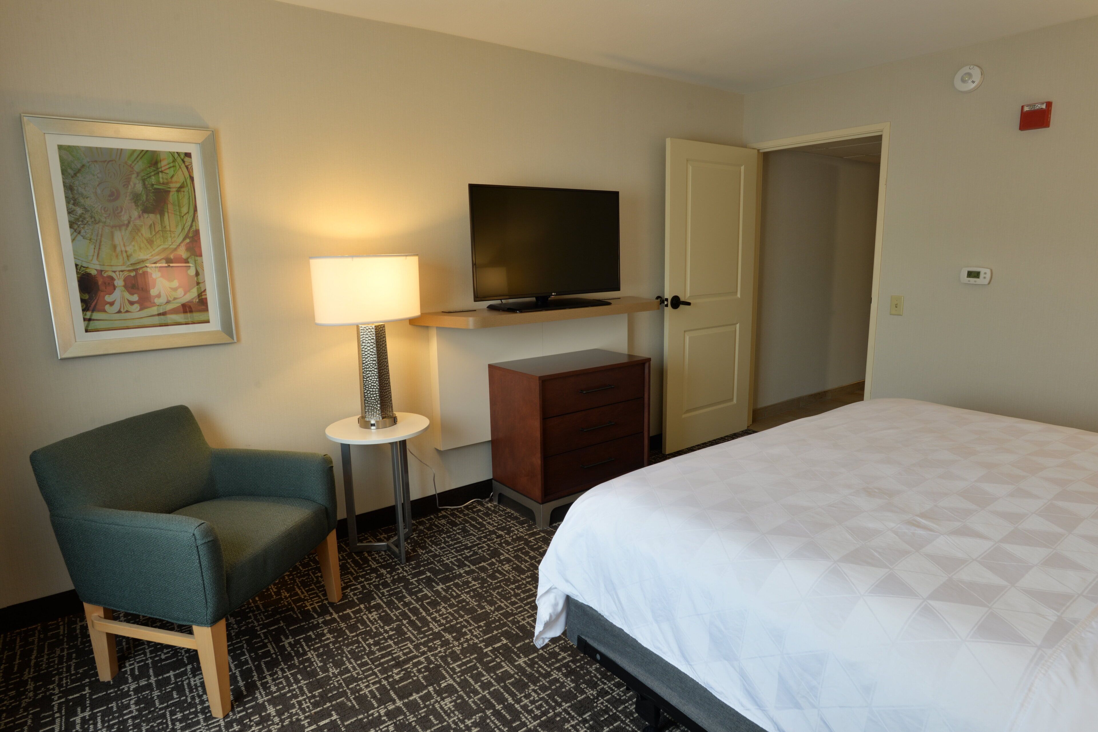 A hotel room with a bed, a chair, a TV on a stand, a lamp, a dresser, and a door to another room.
