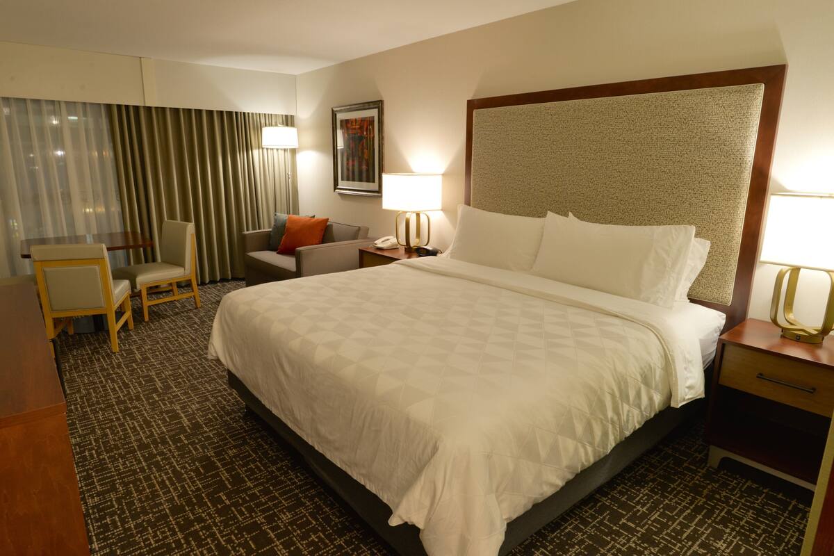 A hotel room with a large bed, a desk, a chair, a nightstand, and a lamp.
