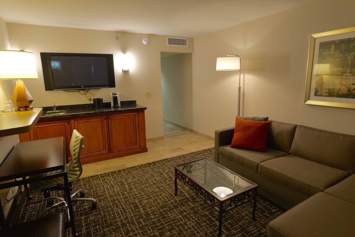 A living room with a sofa, a glass coffee table, a TV mounted on the wall, a desk with a chair, and a kitchen area with cabinets.