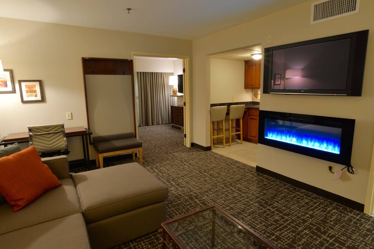 A hotel room with a sofa, ottoman, flat-screen TV, fireplace, and a kitchenette area.