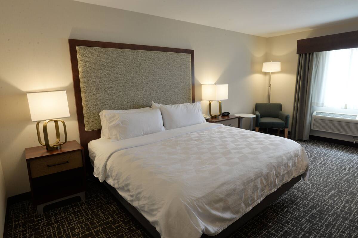 A hotel room with a large bed, two bedside tables with lamps, a chair, and a window with curtains.
