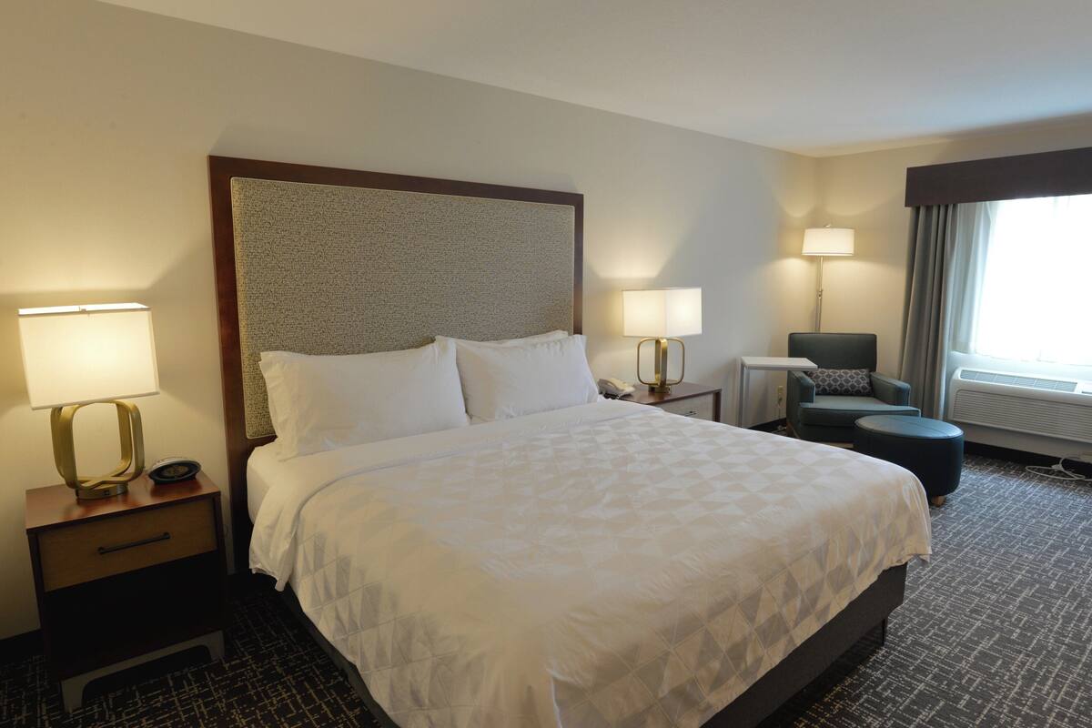 A hotel room with a large bed, two bedside tables with lamps, a chair, and a window with curtains.
