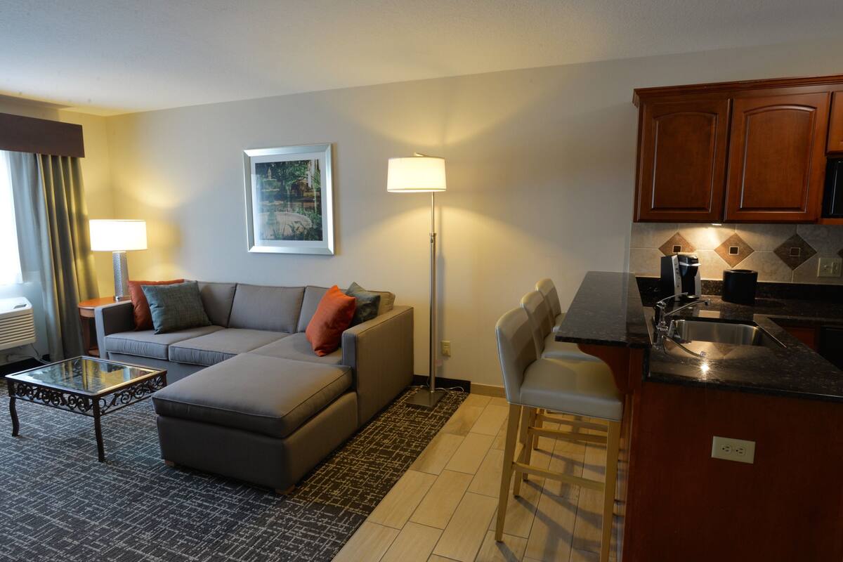 A modern living room with a grey sofa, a glass coffee table, and a floor lamp. There is a kitchen area with wooden cabinets and a sink.
