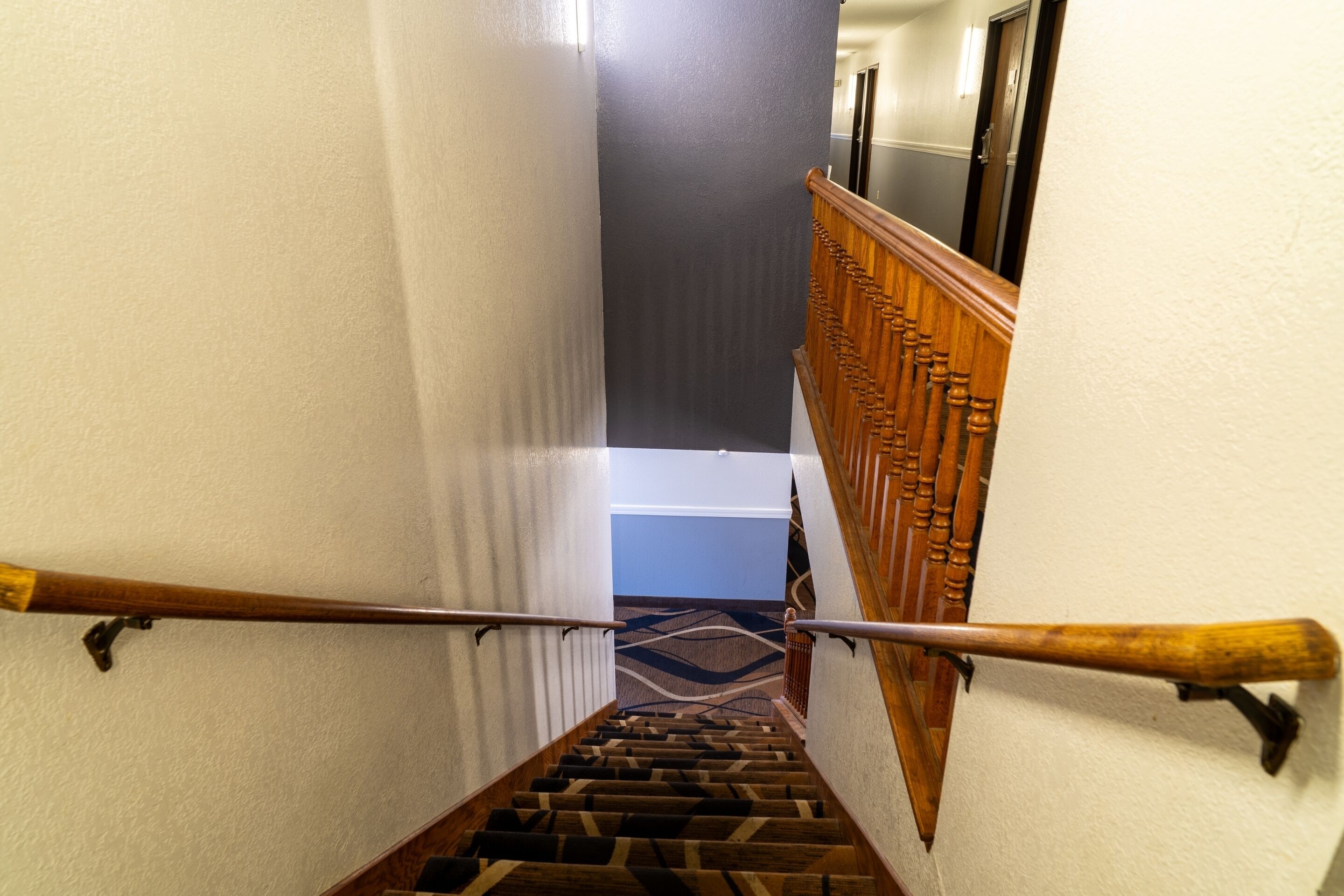 Una escalera con barandilla de madera, peldaños alfombrados y vista a un pasillo con una puerta al final.