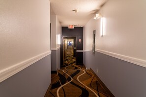 Un pasillo de hotel con piso alfombrado, un letrero de salida montado en la pared y un letrero de prohibido fumar.