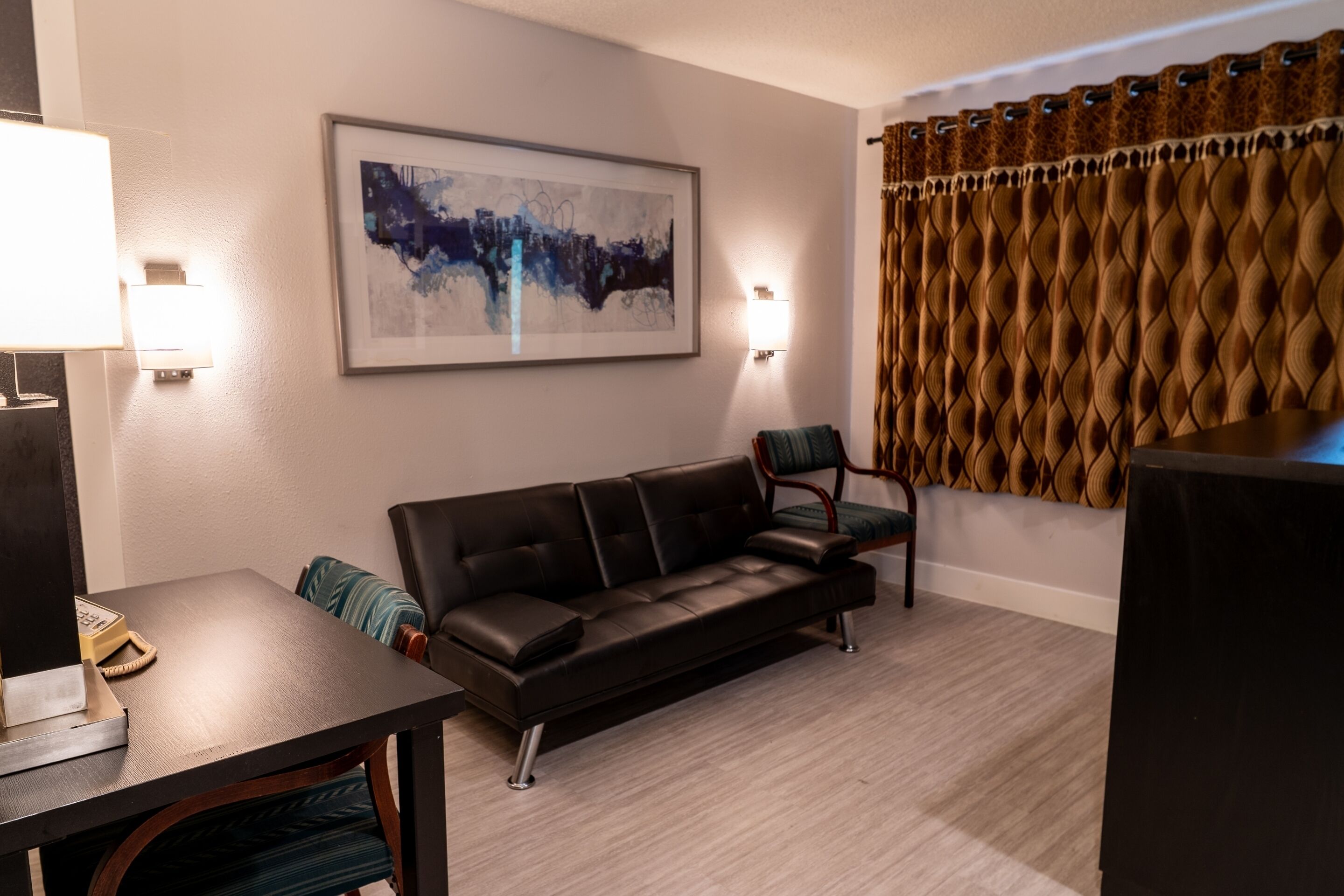 A modern living room with a leather sofa, a dining table with chairs, a framed abstract painting, a wall-mounted light, and a patterned curtain.