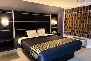 A modern hotel room with a large bed, a headboard with integrated lighting, a bedside table with a lamp, and a patterned curtain.