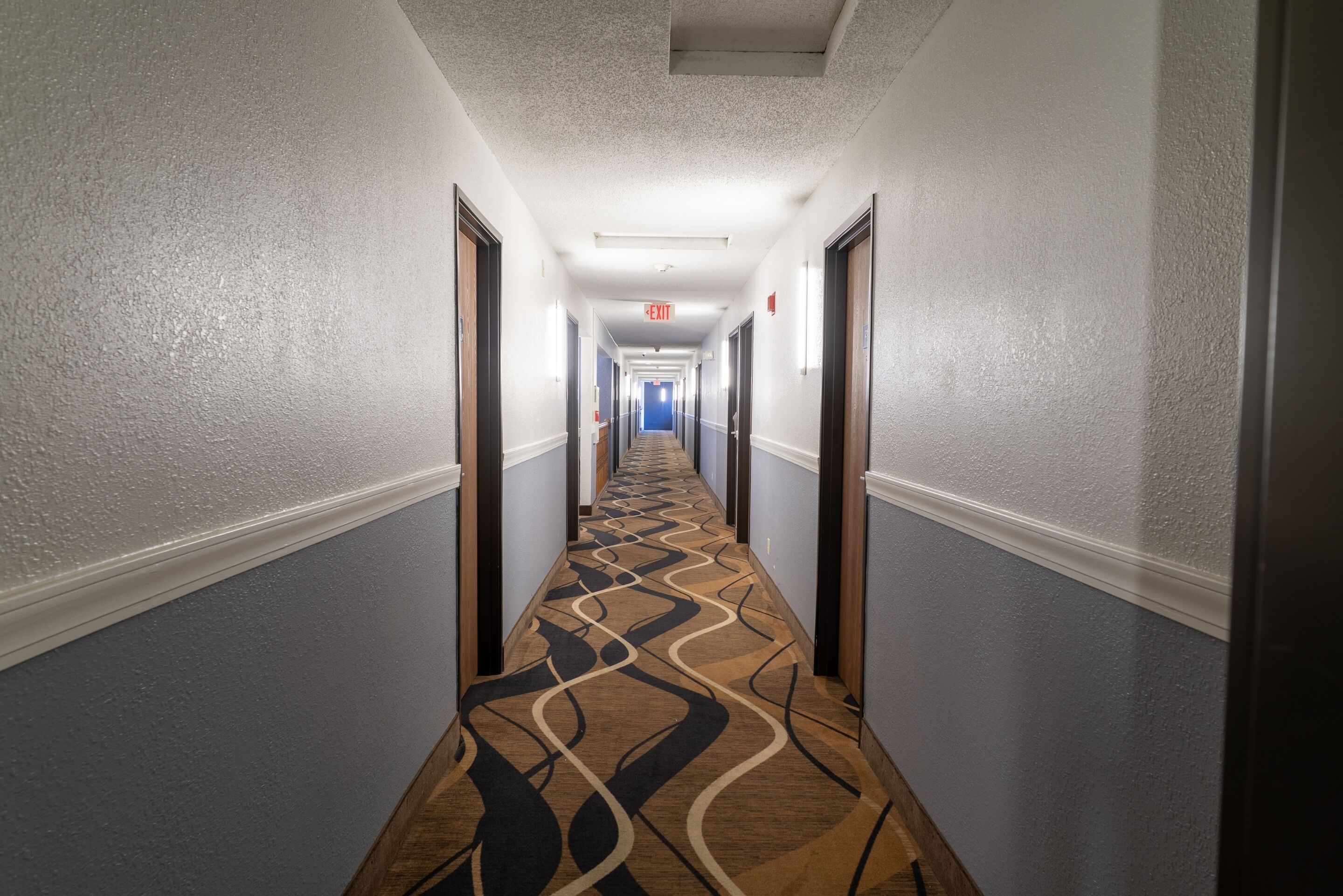 Un corridoio d'albergo con una moquette decorata, porte su entrambi i lati e un segnale di uscita sul soffitto.
