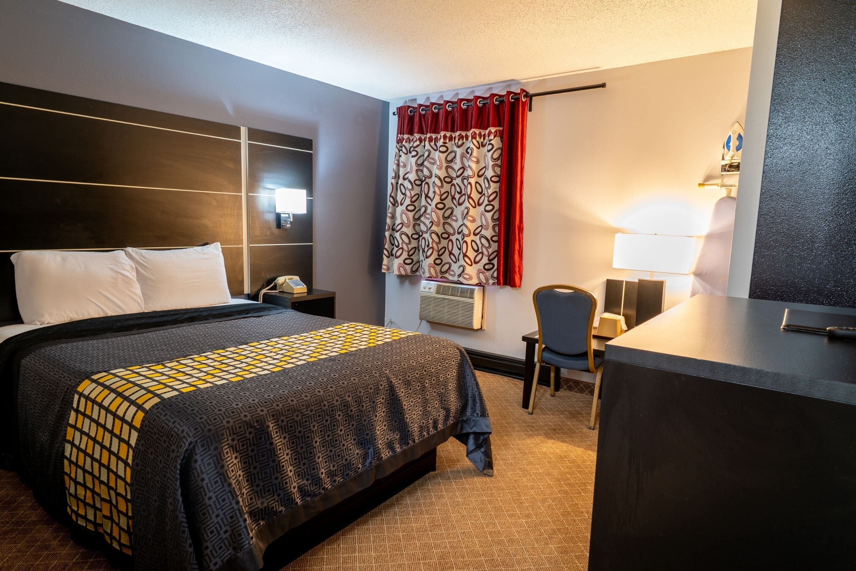 A hotel room with a bed, bedside table, desk, chair, and a desk lamp.