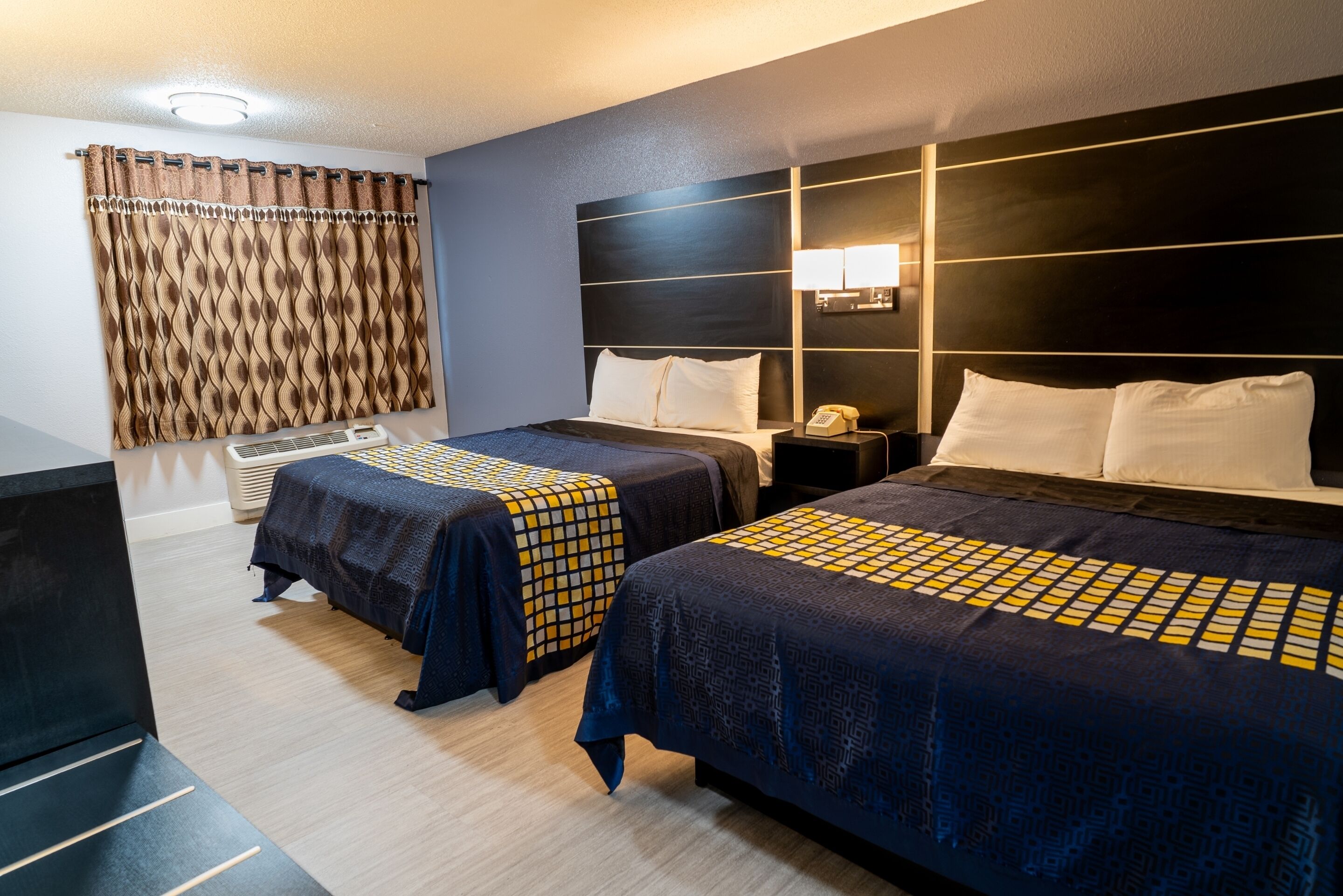 A hotel room with two beds, a bedside table, a lamp, and a heater.
