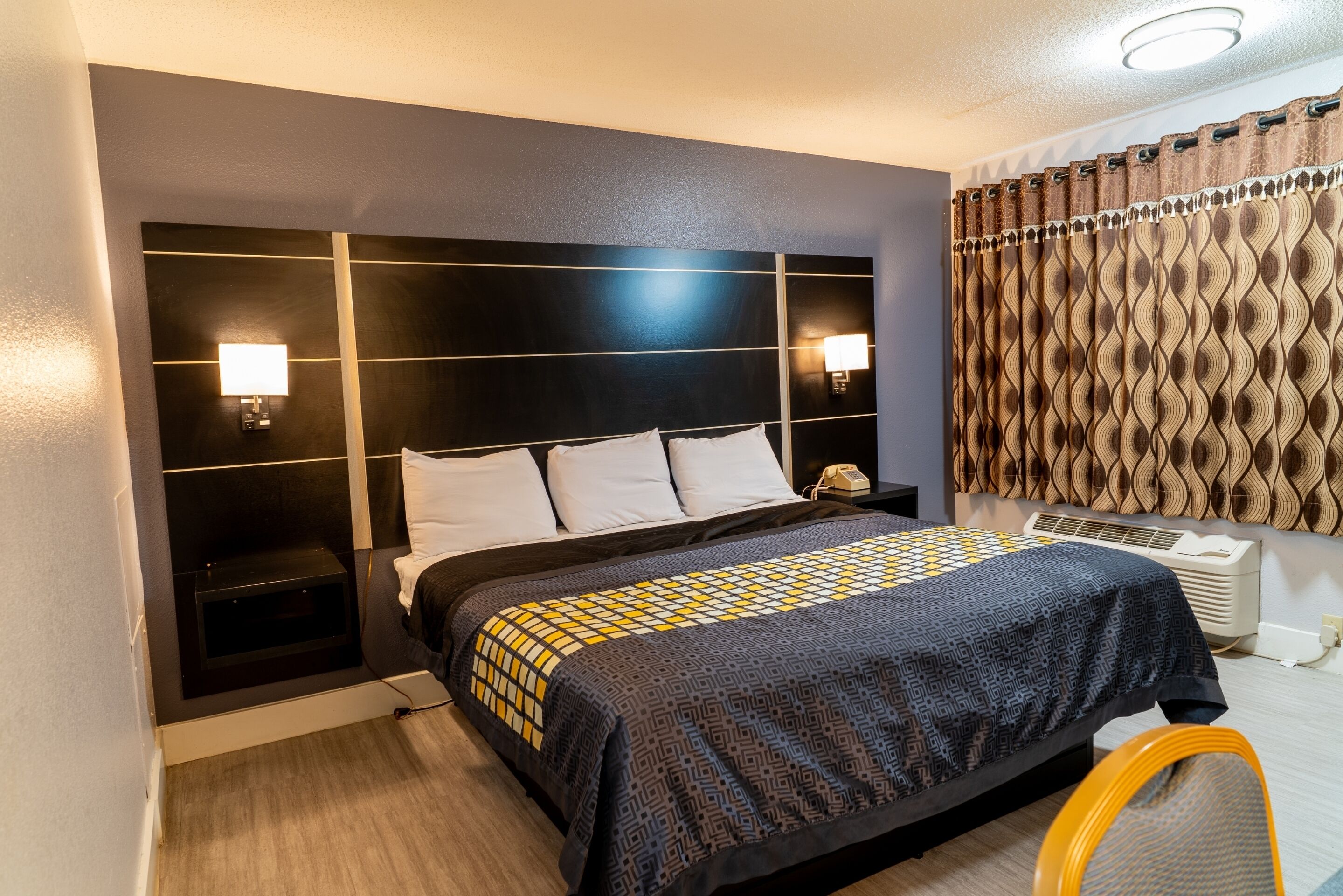 A hotel room with a large bed, bedside lamps, a telephone, a heater, and a patterned curtain.
