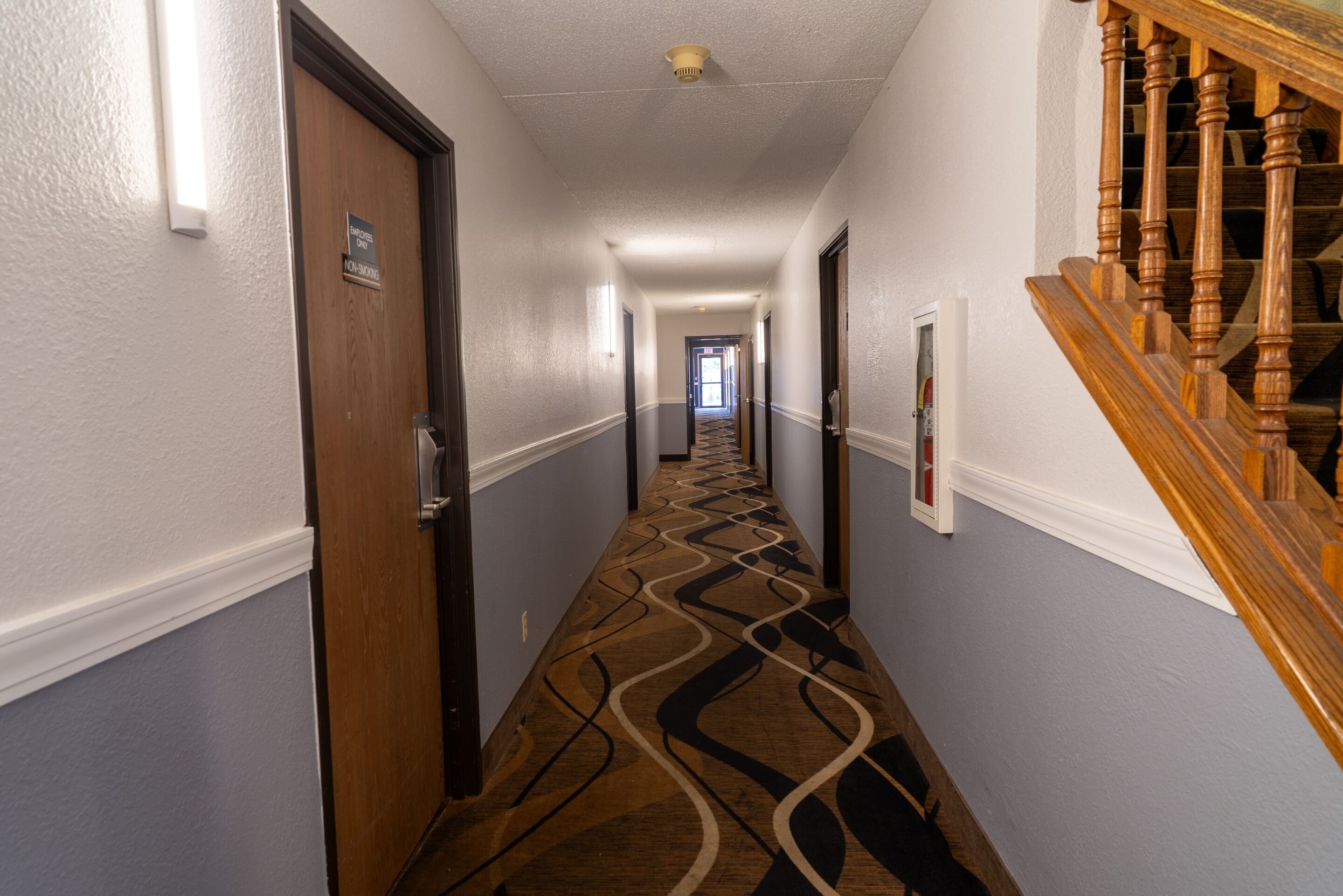 A hotel hallway with a patterned carpet, wooden doors, and a staircase with wooden balusters.