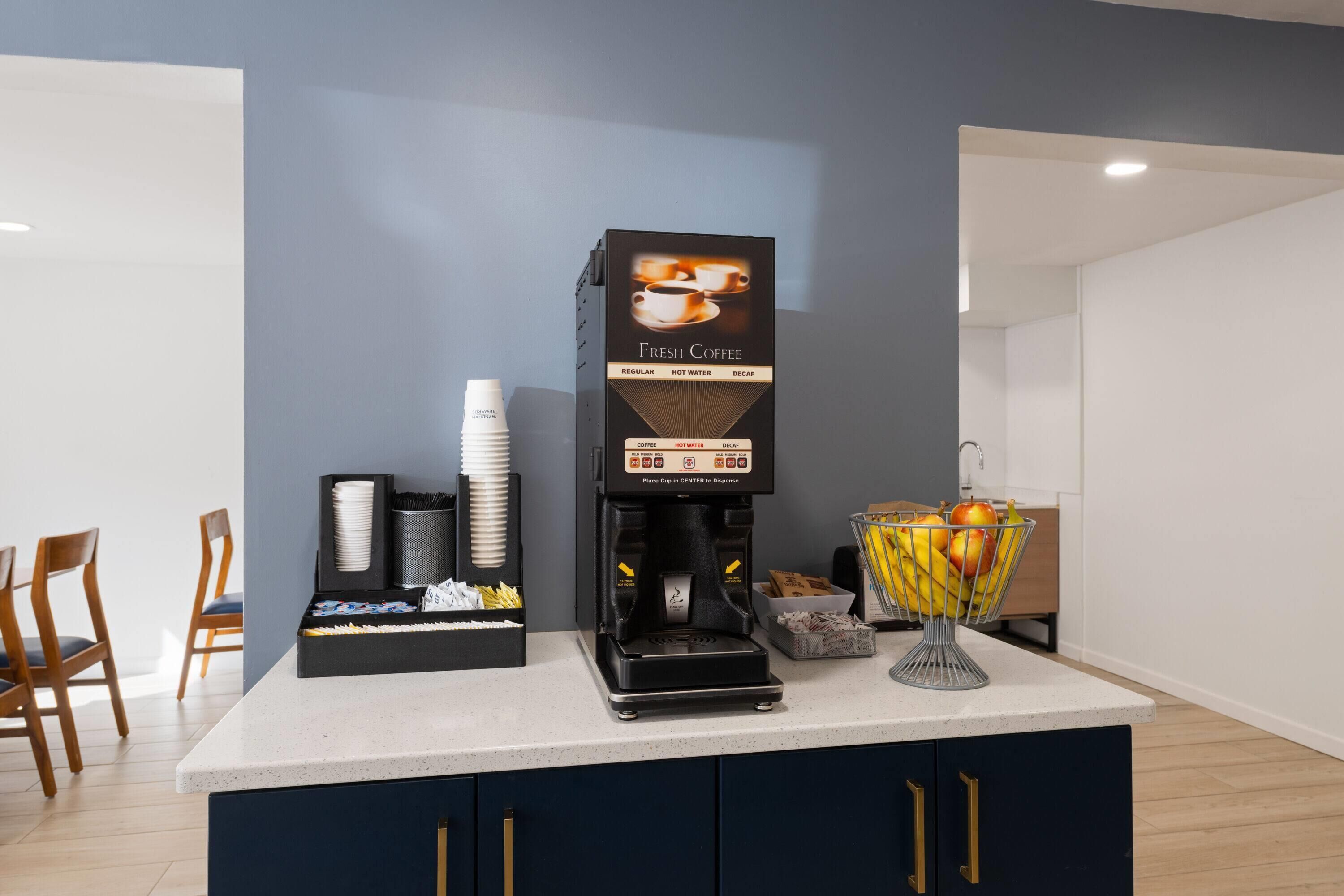 Une machine à café qui propose différentes options : café normal, eau chaude et café décaféiné. Des gobelets jetables et un panier de fruits sont à disposition sur le comptoir.