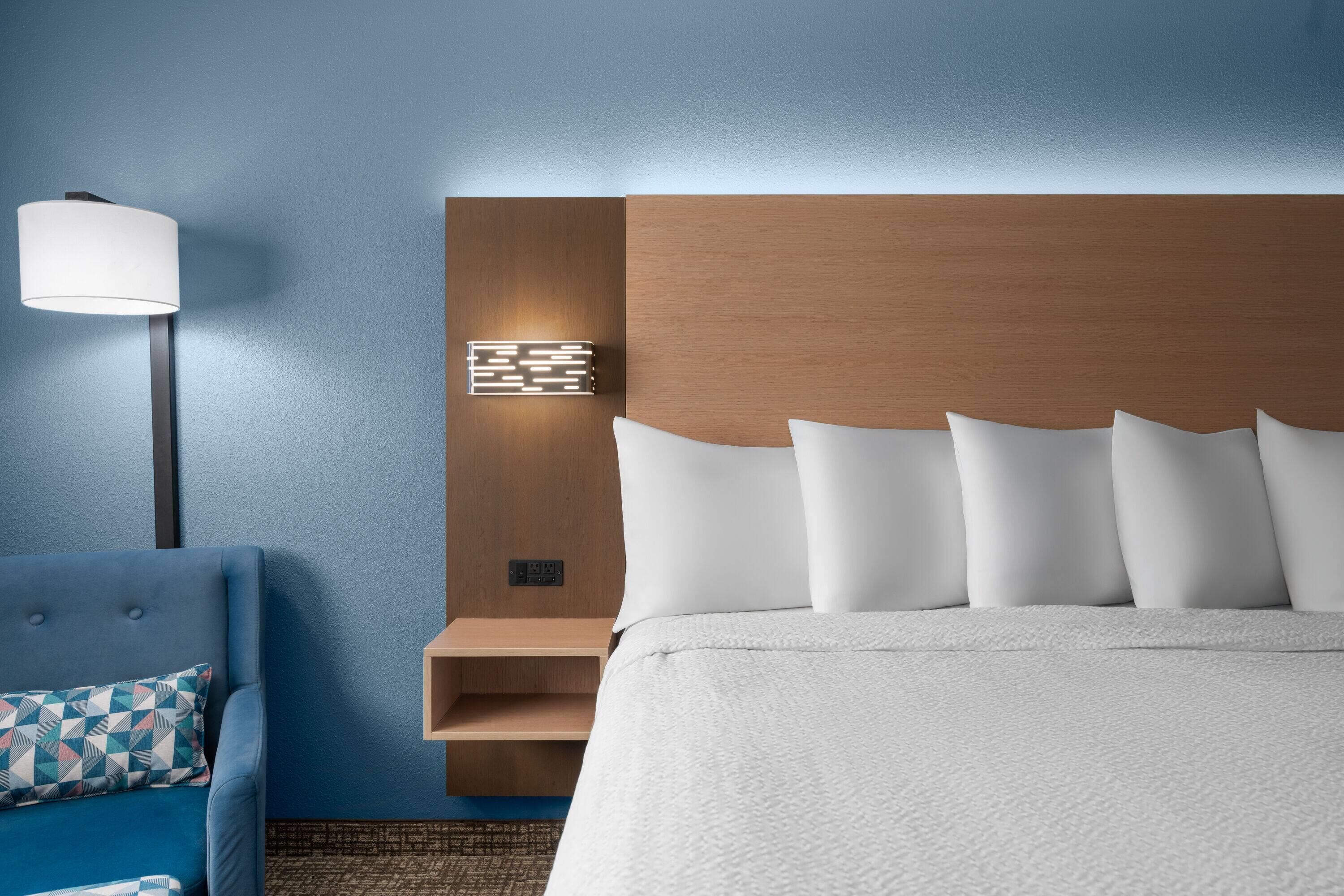 Una habitación de hotel con una cama, un sillón azul, una lámpara de pared y una estantería de madera.