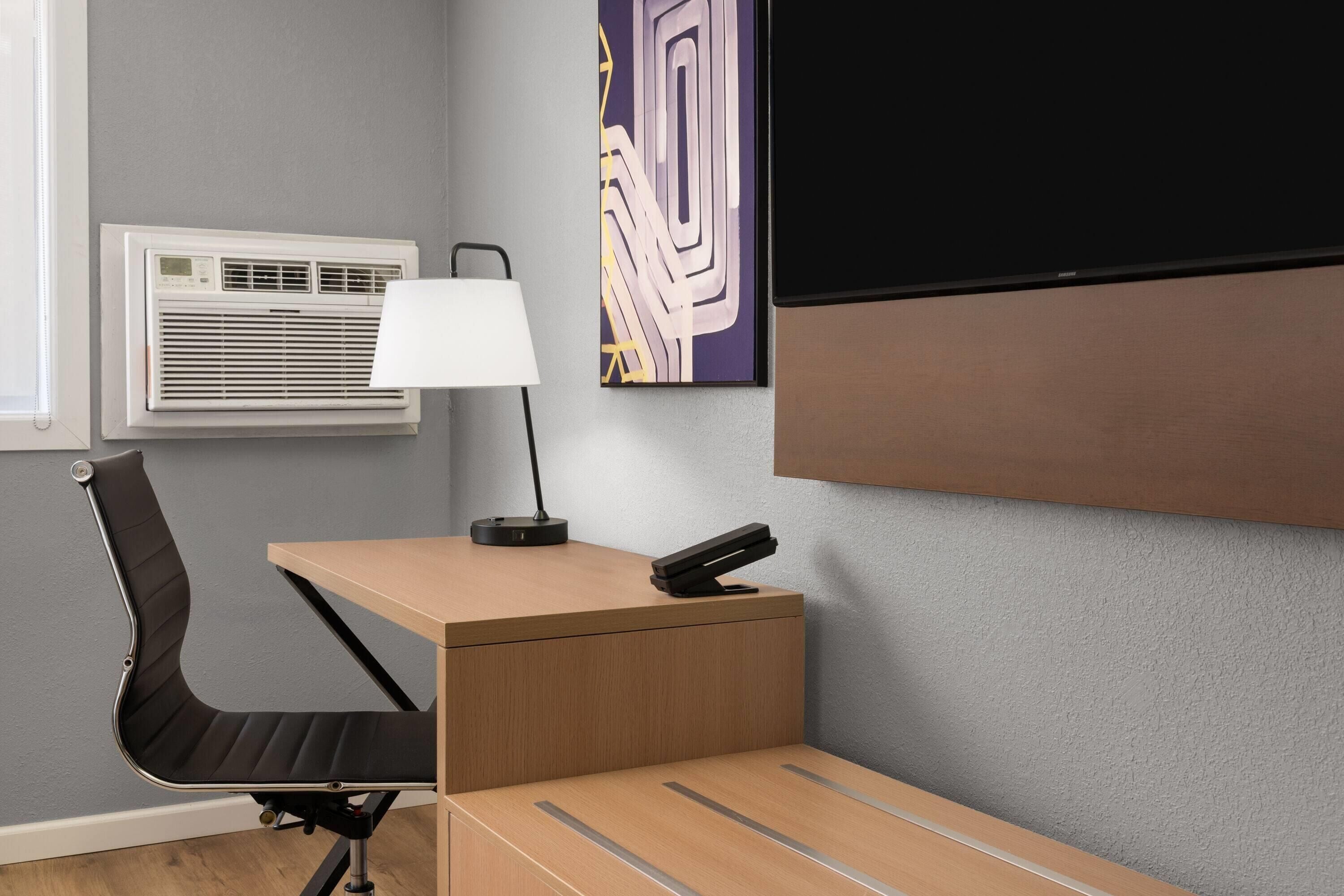Espacio de oficina compacto con escritorio, silla, lámpara, unidad de aire acondicionado y televisión montada.