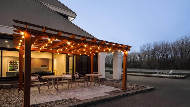 Un área de estar al aire libre techada con mesas y sillas, una pérgola con luces colgantes y vistas a un edificio con fachada de vidrio.