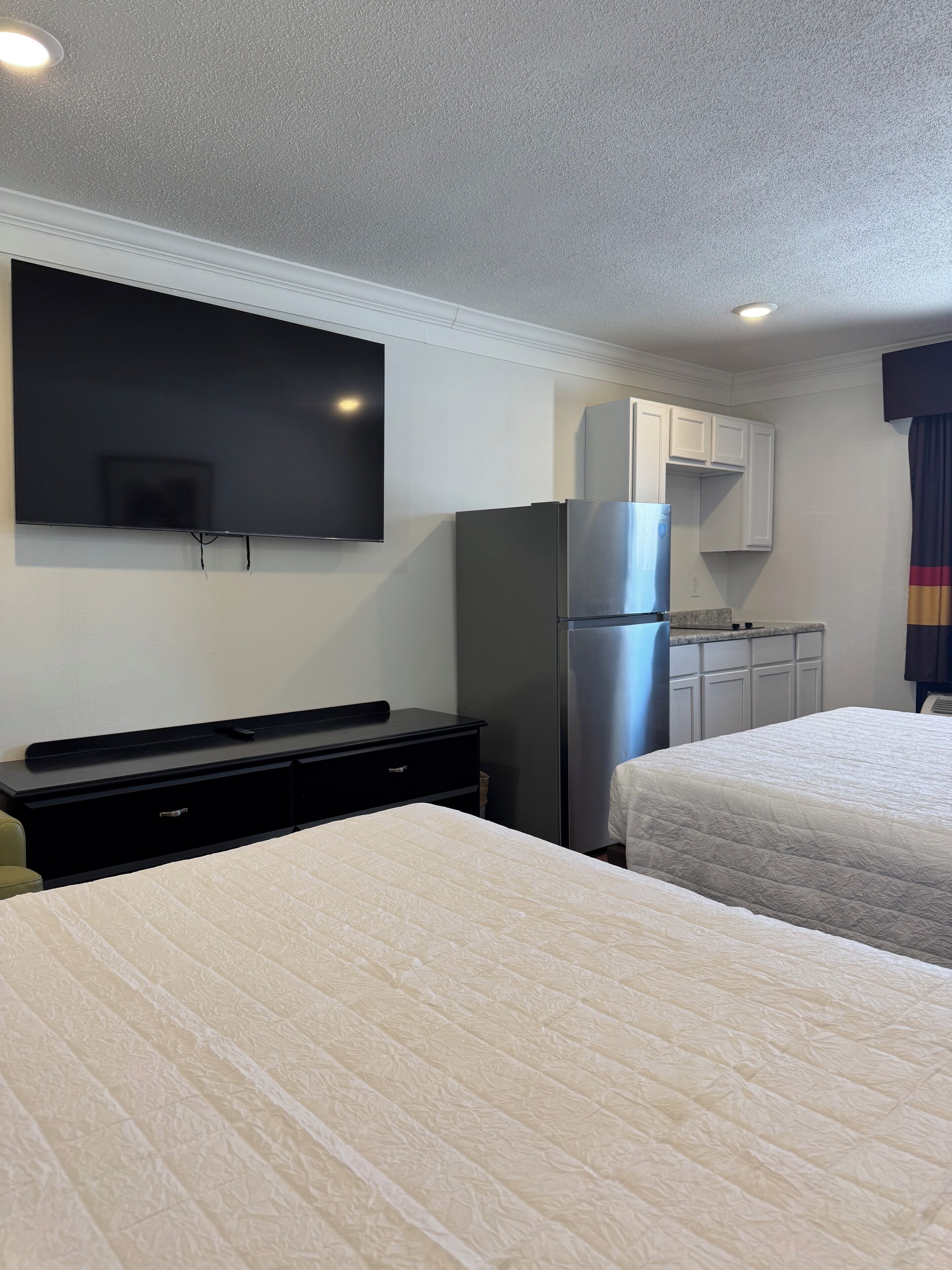 A hotel room with two beds, a flat-screen TV mounted on the wall, a refrigerator, and a small kitchenette area.