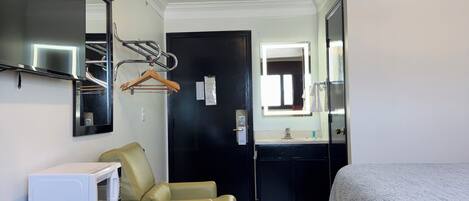 A compact hotel room with a bed, a small desk with a microwave, a chair, a mirror, and a door leading to a bathroom.