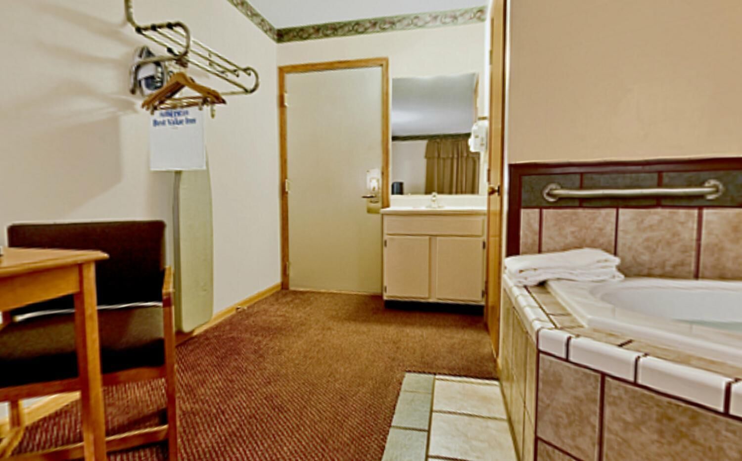 Habitación de hotel con cama, un escritorio pequeño, una silla, baño con lavabo y espejo, y una puerta que da a otra habitación.